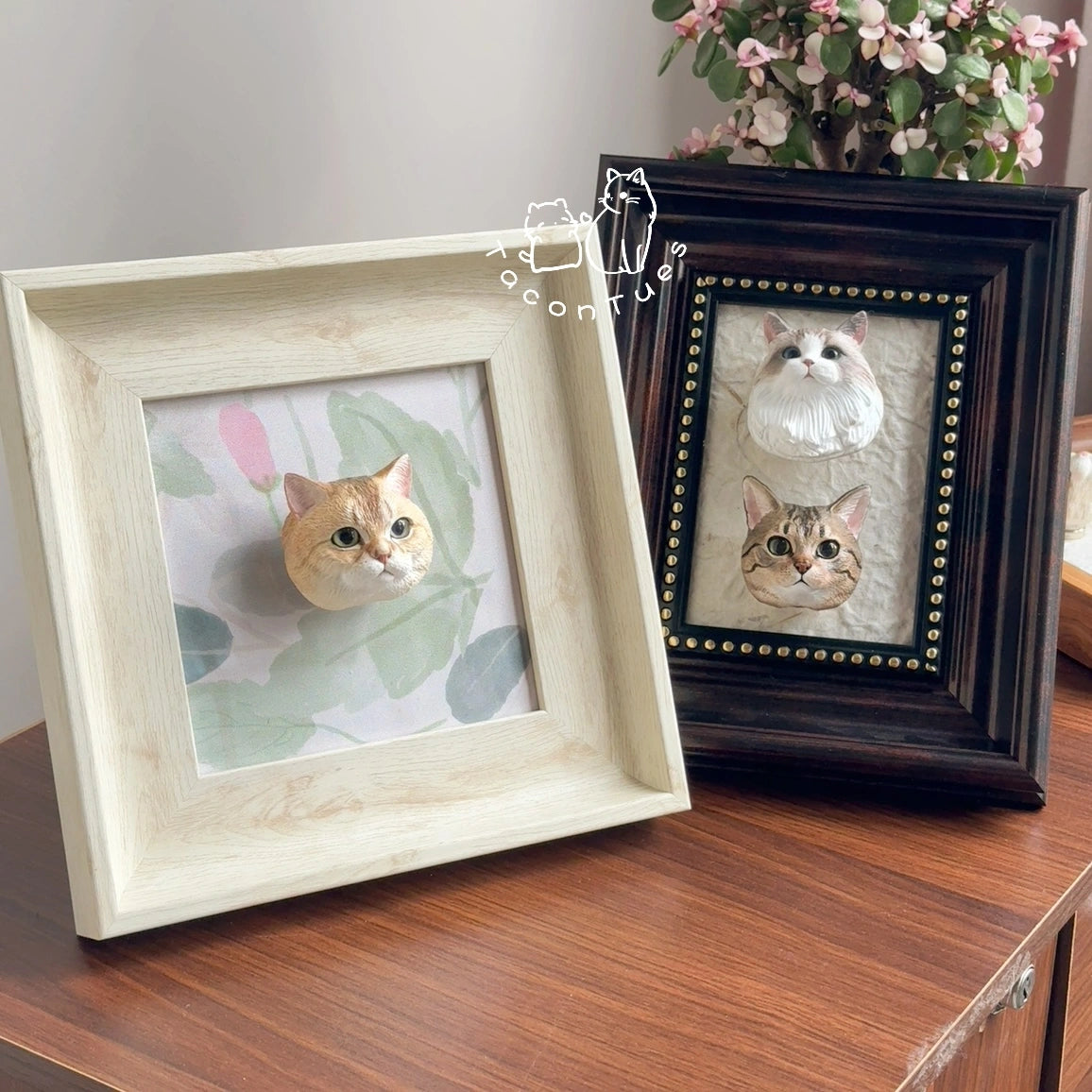 Two custom hand-painted cat figurines in magnetic display frames on a wooden cabinet, one light frame and one dark ornate frame